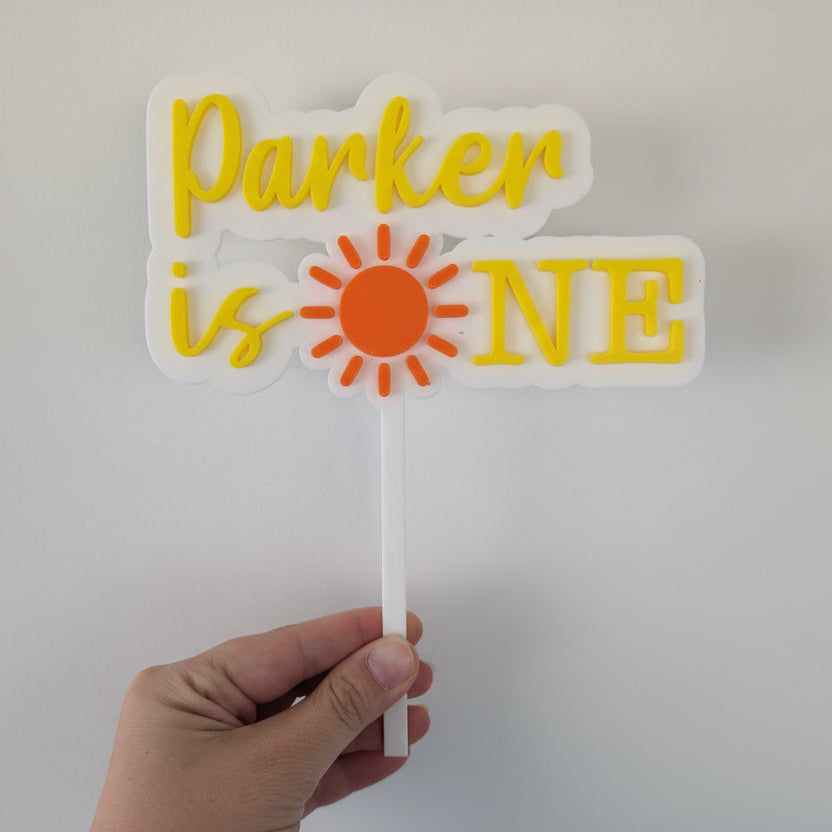 Hand holding a decorative cake topper with 'Parker is ONE' text and sun design.